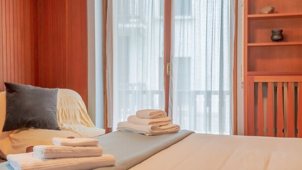 Une chambre d’hôtel comprenant un lit, une table de chevet avec des serviettes, une fenêtre avec des rideaux légers et une étagère en bois ornée d’objets décoratifs.