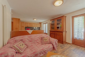Quarto com cama, armários de madeira, área de cozinha e janela com cortinas estampadas com corações.