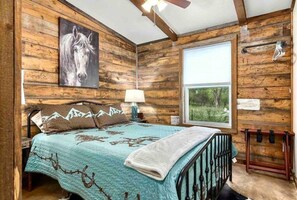 A rustic wooden room with a bed, a window, a horse painting, and a ceiling fan.