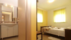 A hotel room with a wooden floor, a bed with a white bedspread, a wooden nightstand, a wooden chair, a small table, a mirror, a sink with a tiled backsplash, and two windows with yellow curtains. - Villa with pool for 6 people (Pula)