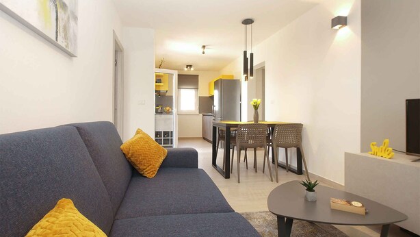 A modern living room with a grey sofa, yellow cushions, a dining area with a table and chairs, and a kitchen in the background. - Holiday home with air conditioning (Fažana)