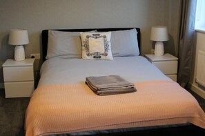A neatly made bed with a folded towel, two bedside lamps, and a decorative pillow. - 1-bed House - Sleeps 2 - Parking, Wifi (Peterborough)