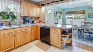 A kitchen with wooden cabinets, a dishwasher, and a coffee maker. There is a dining area with a table and chairs. - 120 Bay Mar Dr (Fort Myers Beach)