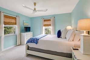 Une chambre à coucher avec un lit, un ventilateur de plafond, un téléviseur posé sur une commode et des fenêtres équipées de stores.