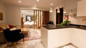A modern kitchen with white cabinets, a black countertop, and pendant lighting. There is a dining area with a black chair and a sofa. - Suite Bali by RentalSevilla (Sevilla)