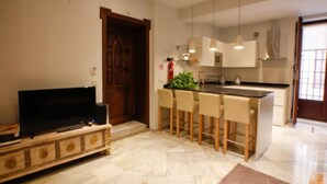 A modern kitchen with a central island, bar stools, and a television on a wooden cabinet. - Suite Bali by RentalSevilla (Sevilla)