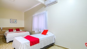 A hotel room with two beds, each with white bedding and red accents, a window with white curtains, and a wall-mounted air conditioner. - Tropical Brisa (São Sebastião)