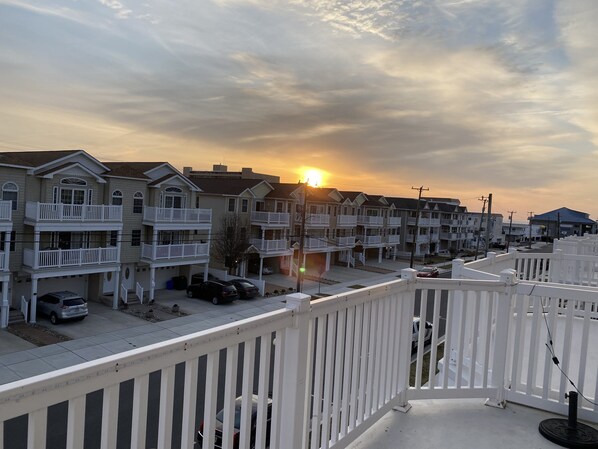 A row of multi-story residential buildings with balconies, a clear sky, and the sun setting in the background.