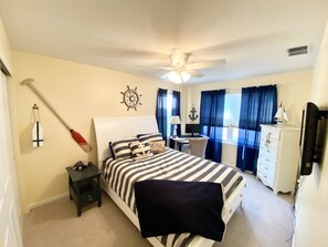 A bedroom with a bed, a desk, a chair, a nightstand, a ceiling fan, and a window with curtains.