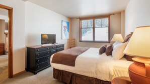 Un dormitorio con cama, cómoda, televisor, ventana con persianas y lámpara.