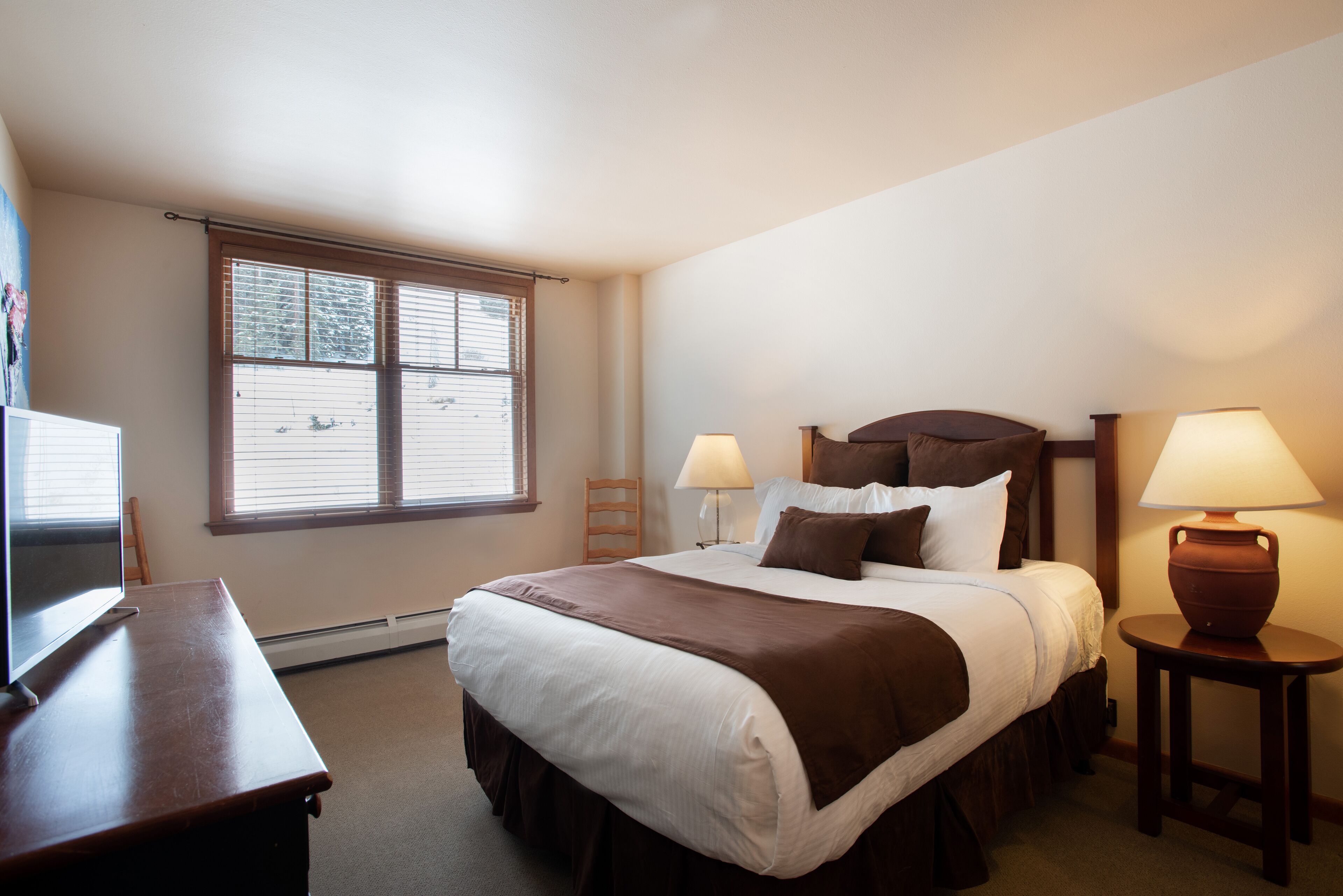 A neatly arranged hotel room with a large bed, two bedside tables with lamps, a desk, and a window with blinds.