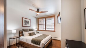 Une chambre avec un lit en bois, un ventilateur de plafond, une fenêtre avec des stores, une table de chevet avec une lampe et un tableau encadré au mur.