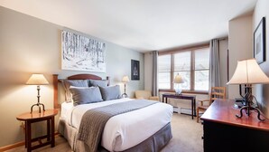 Hotellihuone, jossa on suuri sänky, kaksi yöpöytää valaisimineen, työpöytä, tuoli ja ikkuna, jossa on sälekaihtimet.