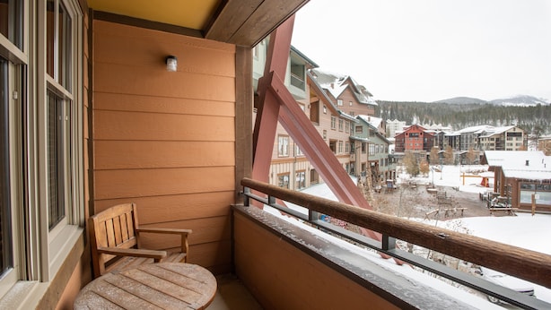 Un balcon avec une table et une chaise en bois, donnant sur une ville enneigée, avec des bâtiments et des montagnes en arrière-plan.