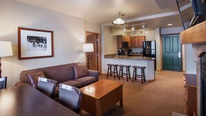 A living room with a sofa, coffee table, and bar stools, a kitchen with a refrigerator, microwave, and stove, and a framed picture on the wall. - Zephyr Mountain Lodge, Condo | 4th Floor Tucked Into The Woods Ski-In/Ski-Out (Select-Rated Condo 1402) (Winter Park)