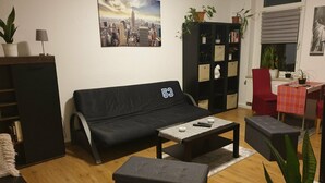 Ein Wohnzimmer mit einer schwarzen Couch, einem Couchtisch und einem roten Sessel. Ein Bücherregal beherbergt Pflanzen und ein Stadtbild.