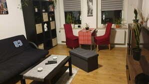 Ein Wohnzimmer mit einem schwarzen Sofa, einem Couchtisch, zwei roten Sesseln, einem kleinen Tisch mit roter Tischdecke und einem Bücherregal.