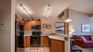 A modern kitchen with wooden cabinets, stainless steel appliances, and a central island. There is a dining area with a red sofa and a lamp. - Zephyr Mountain Lodge, Condo , 2 bedroom (Select-Rated Condo 1604) (Winter Park)