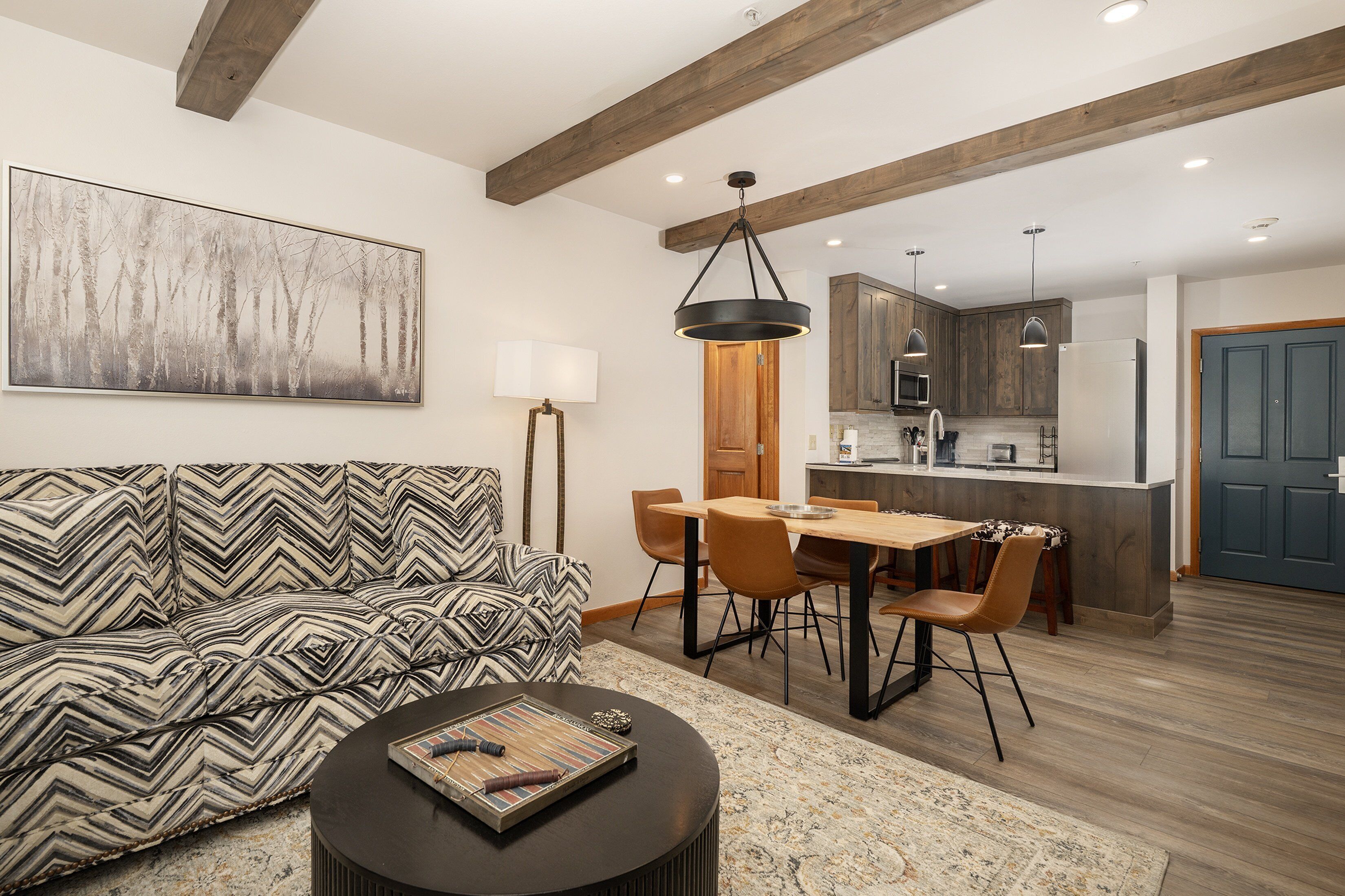 Una sala de estar moderna con un sofá estampado, un comedor con sillas de madera, una cocina con armarios de madera y un cuadro abstracto grande en la pared.