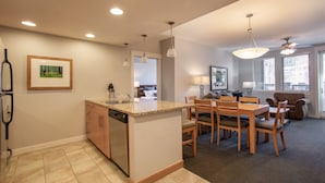 A modern kitchen with a central island, wooden dining table, and chairs. There is a built-in dishwasher and a refrigerator on the left. The room has a ceiling fan and recessed lighting.