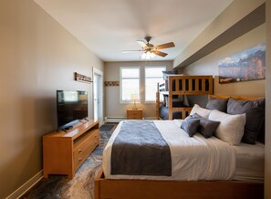A bedroom with a bed, a TV, a ceiling fan, a window, and a painting on the wall.