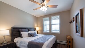 A bedroom with a bed, nightstand, lamp, ceiling fan, windows, and a small wooden stool. - Fraser Crossing/Founders Pointe, Condo , 2 bedroom (Premium-Rated Condo 3506) (Winter Park)