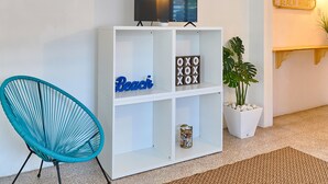 Un salon moderne avec une bibliothèque blanche, un fauteuil bleu, un téléviseur à écran plat et un panneau décoratif représentant une maison de plage accroché au mur.