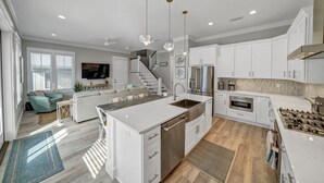 A modern kitchen with white cabinetry, a central island, stainless steel appliances, and a dining area with a sofa and chairs. - Coastal Luxury on 30A: 4br/4ba: golf cart, walk to beach, shopping, & dining!   (Santa Rosa Beach)