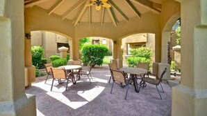 An outdoor patio with tables and chairs, a ceiling fan, and a view of greenery and buildings in the background. - Cozy 1-Bdrm Scottsdale Condo next to pool (Scottsdale)
