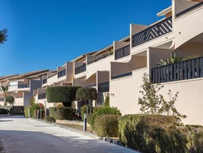 A row of modern, two-story residential buildings with balconies, neatly trimmed hedges, and a clear blue sky.