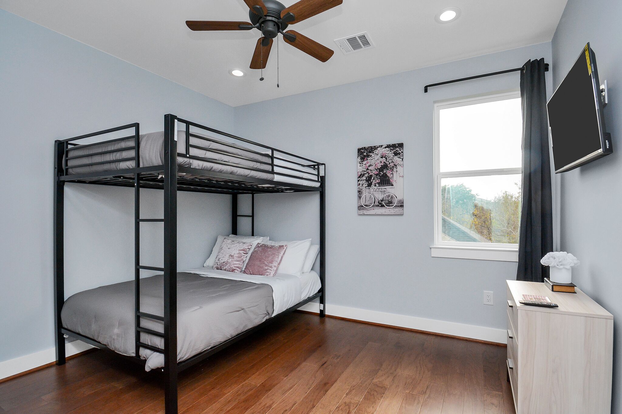 A bunk bed in a room with a ceiling fan, a window with curtains, and a mounted TV.