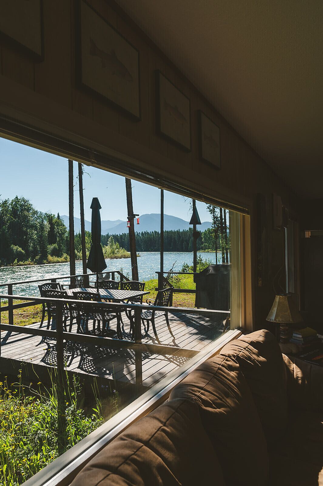 A room with a large window overlooking a lake, a deck area with outdoor furniture, and a cozy seating area with a lamp.