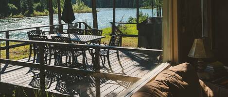 A room with a large window overlooking a lake, a deck area with outdoor furniture, and a cozy seating area with a lamp.
