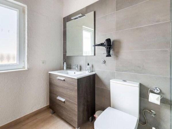 Une salle de bain avec un lavabo blanc, un meuble en bois foncé et des toilettes blanches. Un miroir est placé au-dessus du lavabo et un sèche-cheveux est fixé au mur.