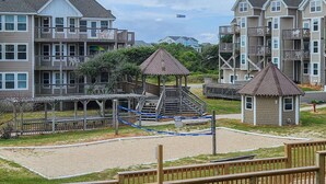 A playground with a slide and a small gazebo, surrounded by a wooden fence and a sandy area. - IT'S A SHORE THING ☀️3BR Oceanfront Condo in Duck w/Community Pool☀️ (Duck)