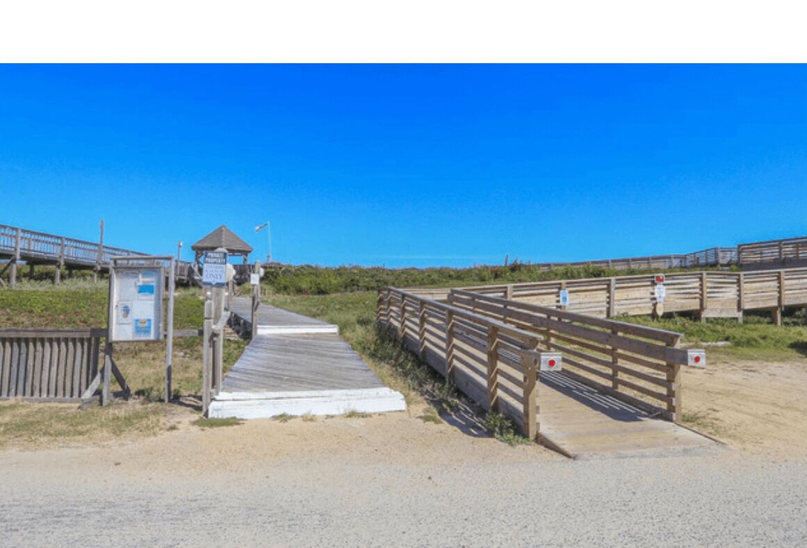 Una passera de fusta que condueix a una zona de platja amb un petit pavelló i una caseta de socorristes.