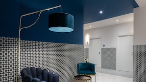 Eine moderne Lobby mit blauer Wand, gemusterten Fliesen, einem blauen Sessel, einer goldenen Stehlampe und einem kleinen runden Tisch.