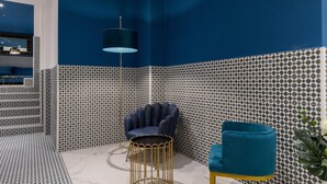 Ein Zimmer mit blauer Akzentwand, strukturiertem Wandpapier, einem runden goldenen Tisch, einem blauen Sessel und einer Stehlampe.