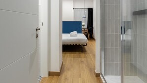 Habitación de hotel moderna con piso de madera, cama con ropa blanca, un escritorio pequeño con una silla y zona de ducha con puertas de vidrio.