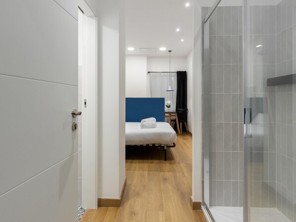 A modern hotel room with a wooden floor, a bed with white linens, a small desk with a chair, and a shower area with glass doors. - Hotelito Boutique Valencia Estación 5 (Valencia)