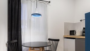 Een kleine keuken met een magnetron, koffiezetapparaat en een spoelbak. Er staat een rond tafel met twee stoelen en een hangende lamp.