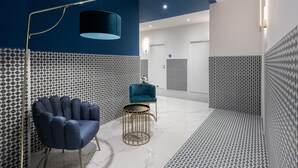 Een moderne hotellobby met behang, een blauwe fauteuil, een gouden vloerlamp en een klein rond tafeltje.