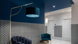 Un vestíbulo moderno con una pared azul oscuro, baldosas estampadas, un sillón azul, una lámpara de pie dorada y una pequeña mesa redonda.
