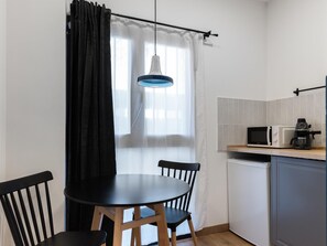 Une cuisine compacte avec une table ronde, deux chaises, un four à micro-ondes, une machine à café et une fenêtre ornée de rideaux légers.