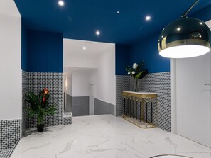 A modern bathroom with a white marble floor, blue and black patterned walls, a gold vanity, and a large black pendant light.
