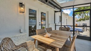 Eine Terrasse mit Holztisch und Korbstühlen, einer weißen Backsteinwand und einer Glastür, die zu einem überdachten Außenbereich führt.