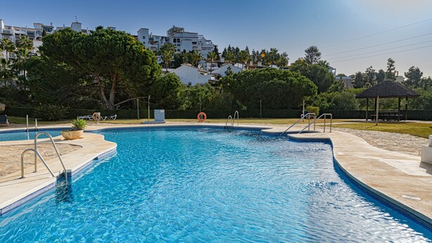 A swimming pool with clear blue water, surrounded by a tiled deck and a grassy area with trees and a gazebo. - ARTE DE LUZ - Fantastic apartment next to the golf course and with a shared pool in Calaho (Mijas)
