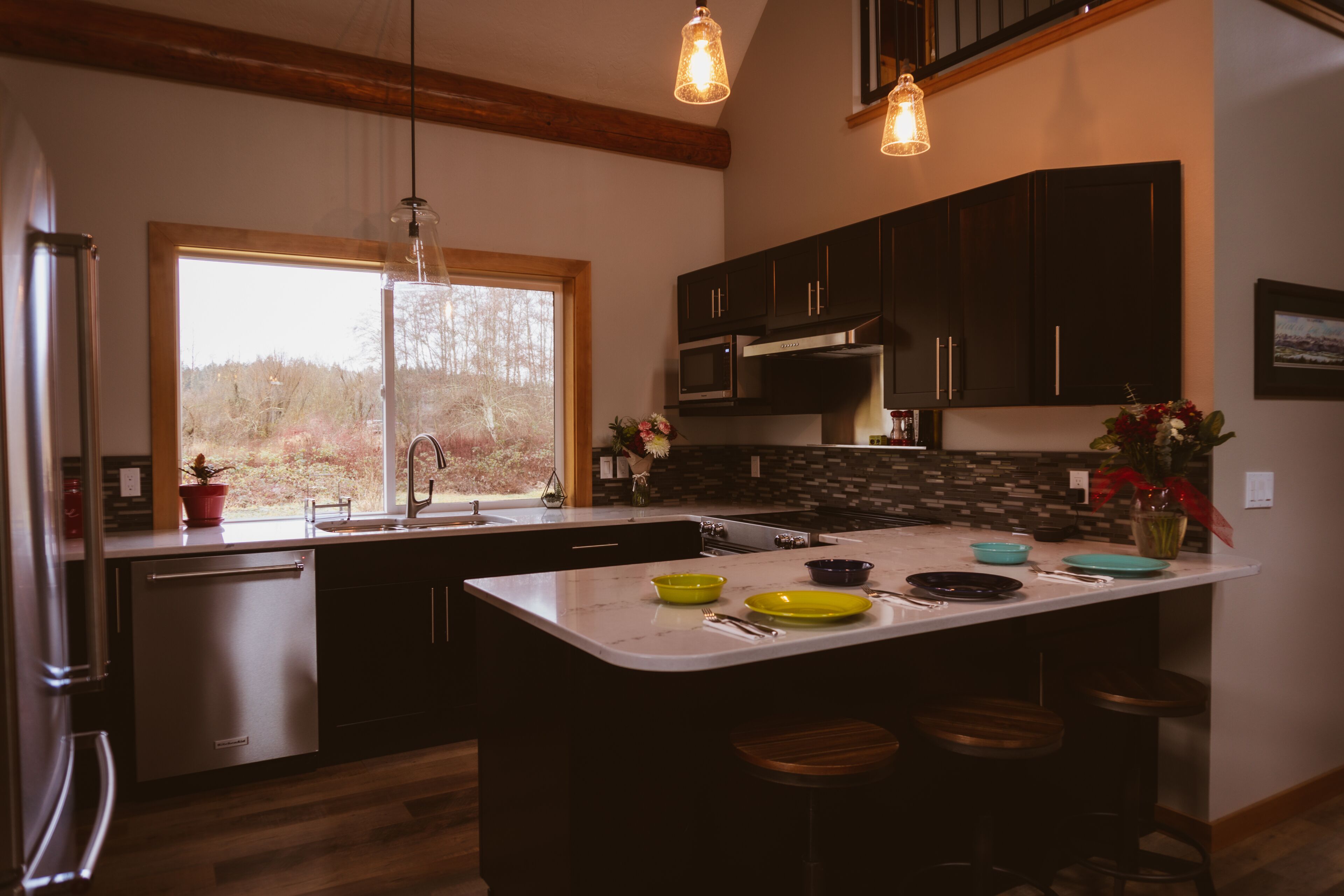 A modern kitchen with dark cabinets, a large island with stools, a built-in oven, and a window with a view of trees.