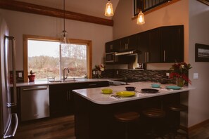 A modern kitchen with dark cabinets, a large island with stools, a built-in oven, and a window with a view of trees.