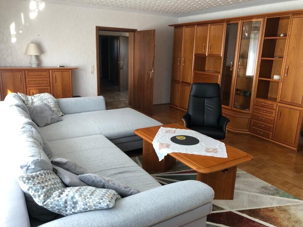 Ein Wohnzimmer mit einer Couch, einem schwarzen Sessel, einem Holzschrank und einem kleinen Tisch mit Platzdeckchen.